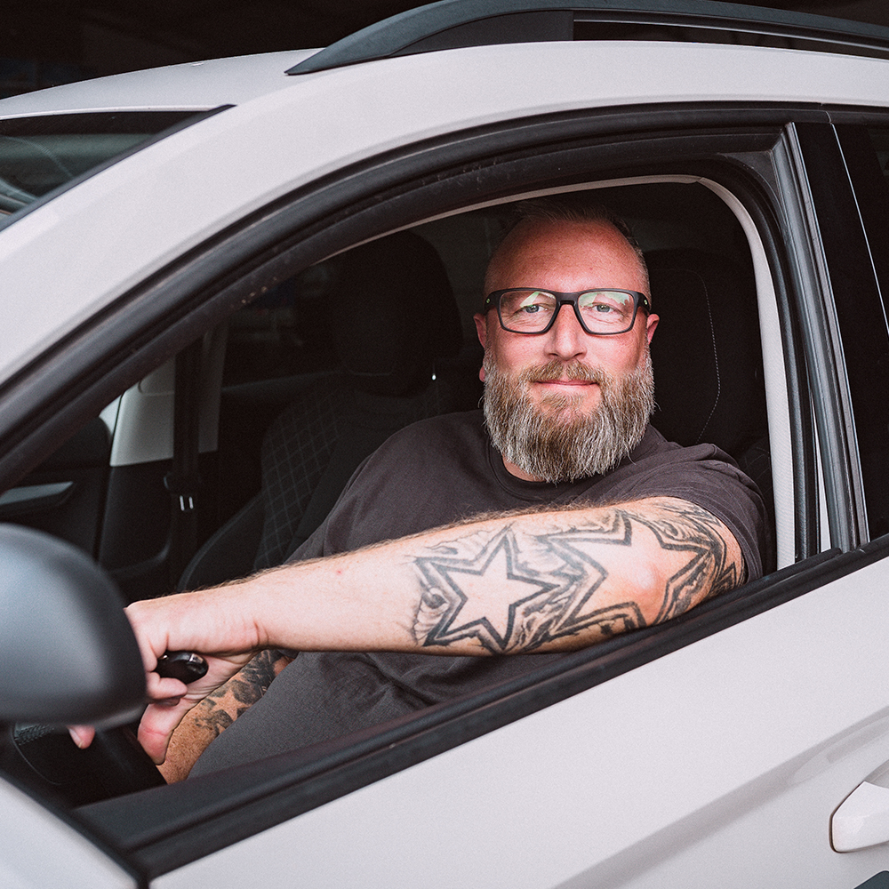 Alexander Veit mit Brille und Tätowierungen auf dem Arm sitzt lächelnd am Steuer eines weißen Fahrschulautos der Fahrschule Hüsich und hält einen Autoschlüssel in der Hand.