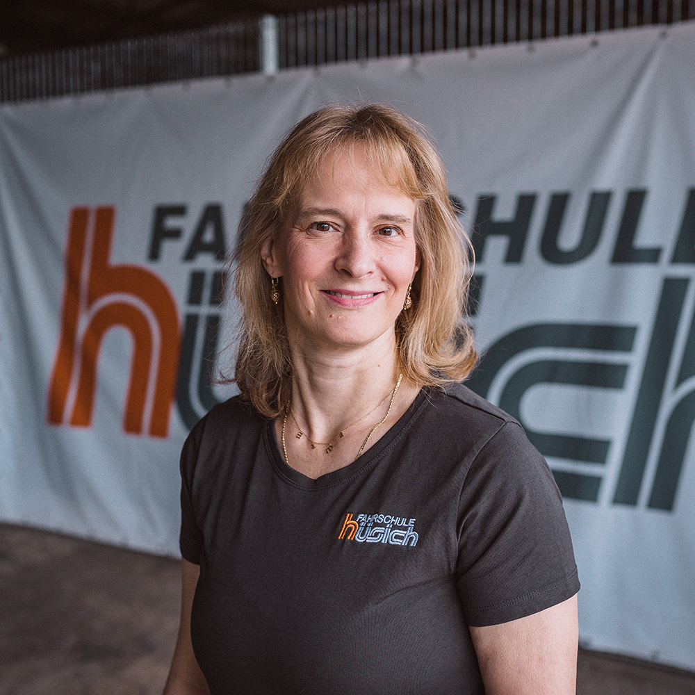 Cathrin Siafakas mit schulterlangem blondem Haar steht vor einem Banner mit dem Logo der Fahrschule Hüsich. Sie trägt ein dunkelgraues T-Shirt mit dem aufgedruckten Logo der Fahrschule und lächelt freundlich in die Kamera.