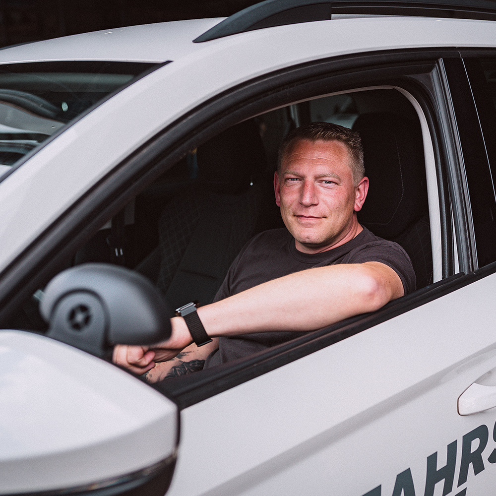 Sebastian Veit lächelt mit kurzgeschorenen Haaren und einer schwarzen Uhr am Handgelenk sitzt am Steuer eines weißen Fahrschulautos der Fahrschule Hüsich und schaut direkt in die Kamera.