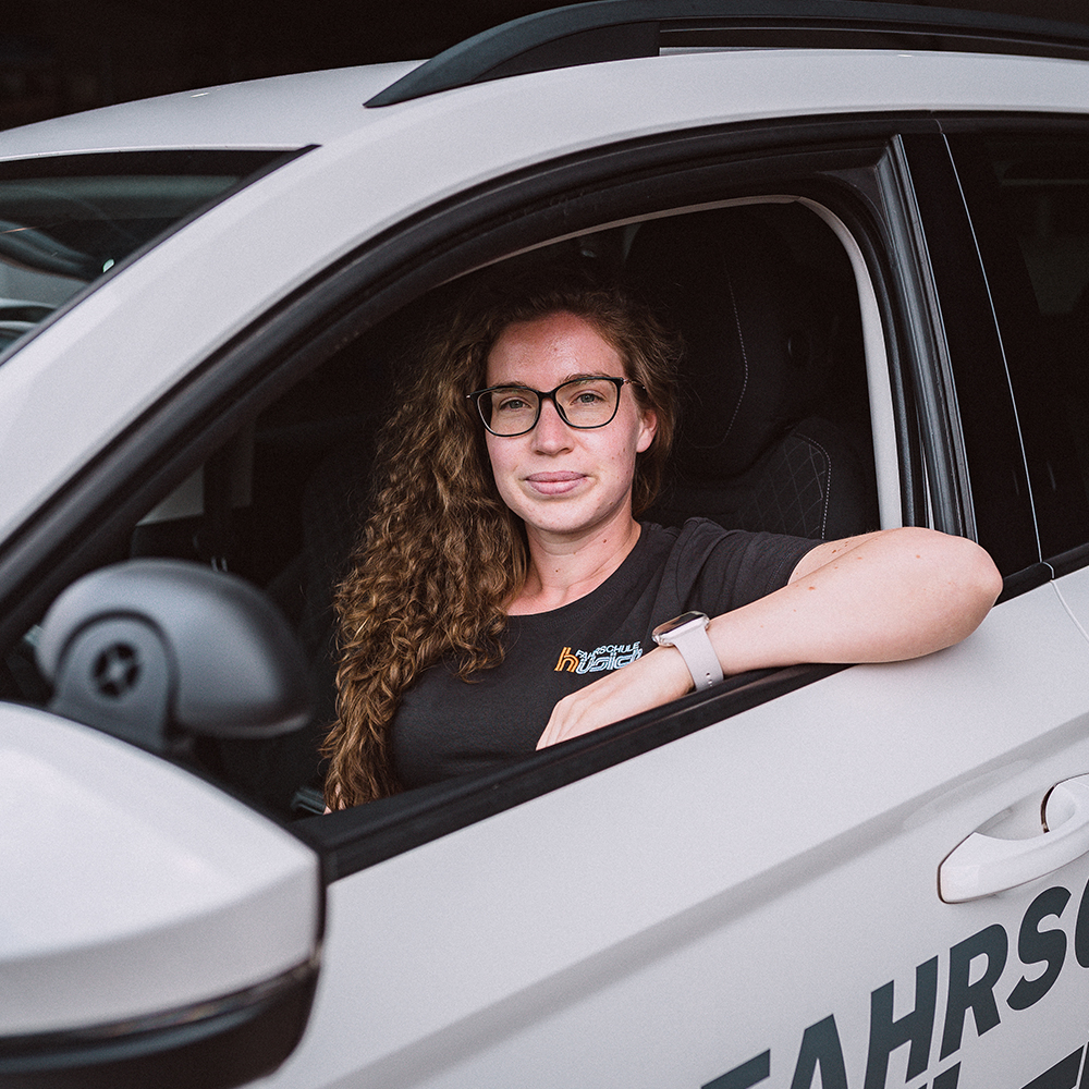 Eine junge Frau mit langen lockigen Haaren und Brille sitzt am Steuer eines weißen Fahrschulautos der Fahrschule Hüsich und schaut in die Kamera.