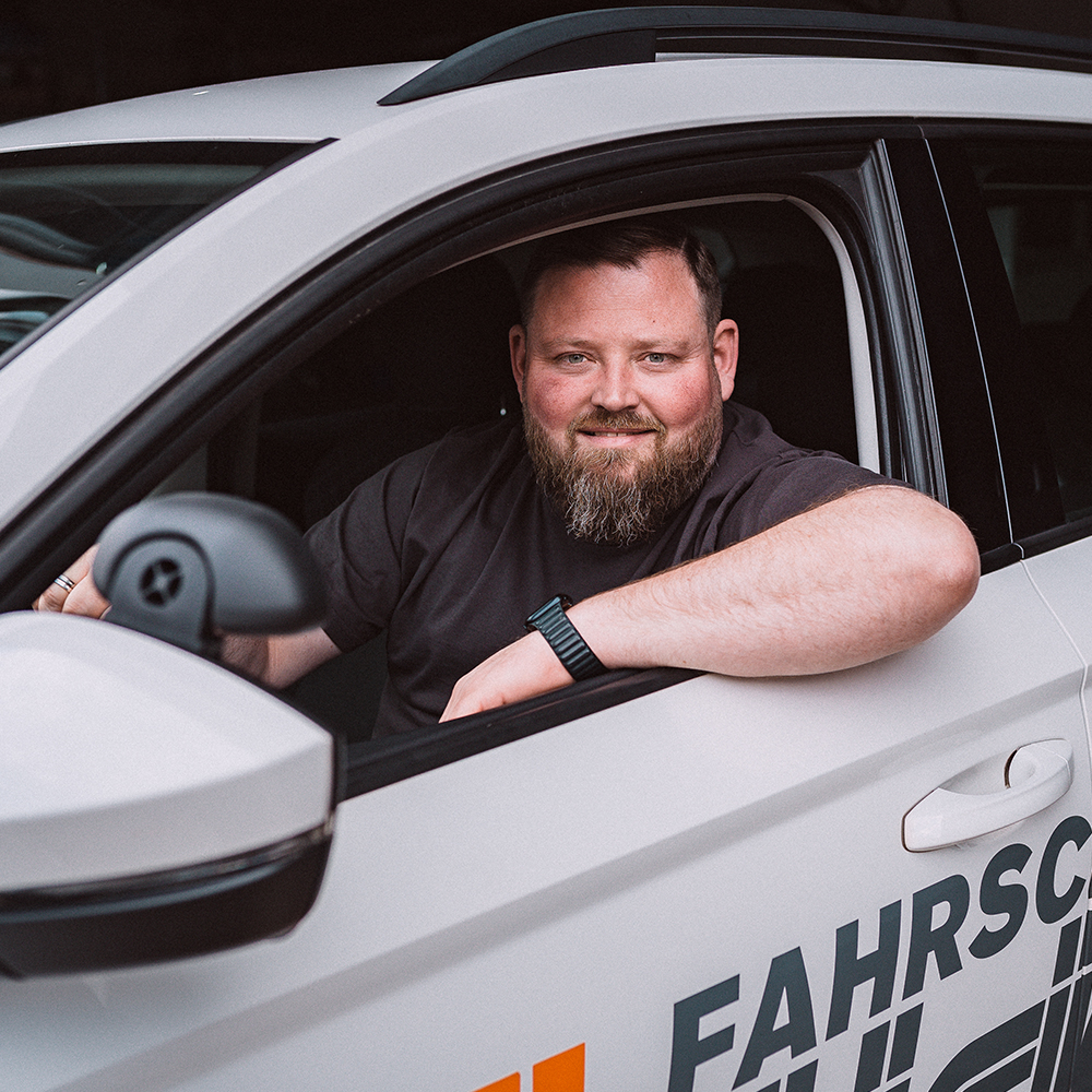 Florian Marotzke mit kurzem Haar und Bart sitzt lächelnd am Steuer eines weißen Autos der Fahrschule Hüsich. Er lehnt den linken Arm entspannt aus dem geöffneten Fenster. Auf der Fahrzeugtür ist das Logo der Fahrschule sichtbar.