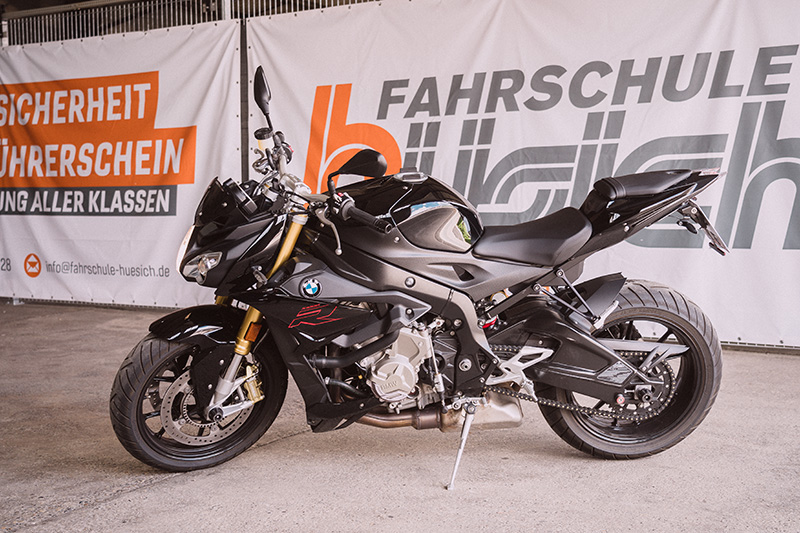 Ein glänzend schwarzes BMW Motorrad steht vor dem orange-weißen Werbebanner der Fahrschule Hüsich.