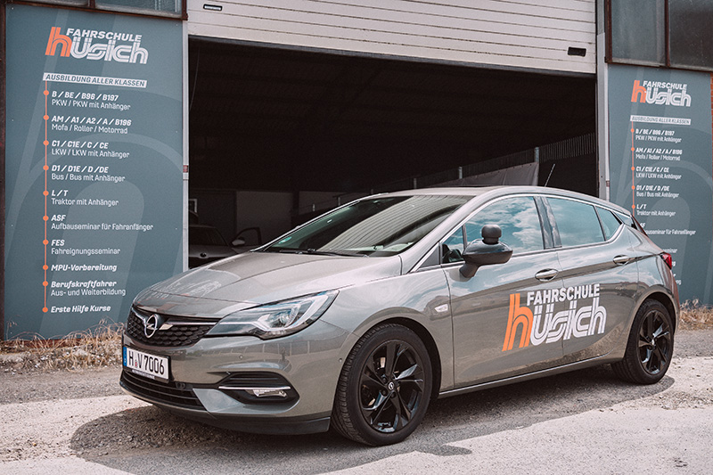 Ein grauer Opel Astra mit dem großen orangen Logo der Fahrschule Hüsich auf der Seite steht vor dem Eingang, neben dem eine Übersicht aller angebotenen Führerscheinklassen angebracht ist.