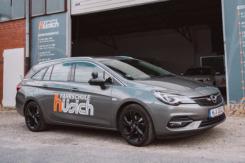 Ein grauer Opel Astra Kombi mit dem Logo der Fahrschule Hüsich steht vor der Einfahrt des Gebäudes, im Hintergrund ist ein weiteres Fahrschulauto erkennbar.