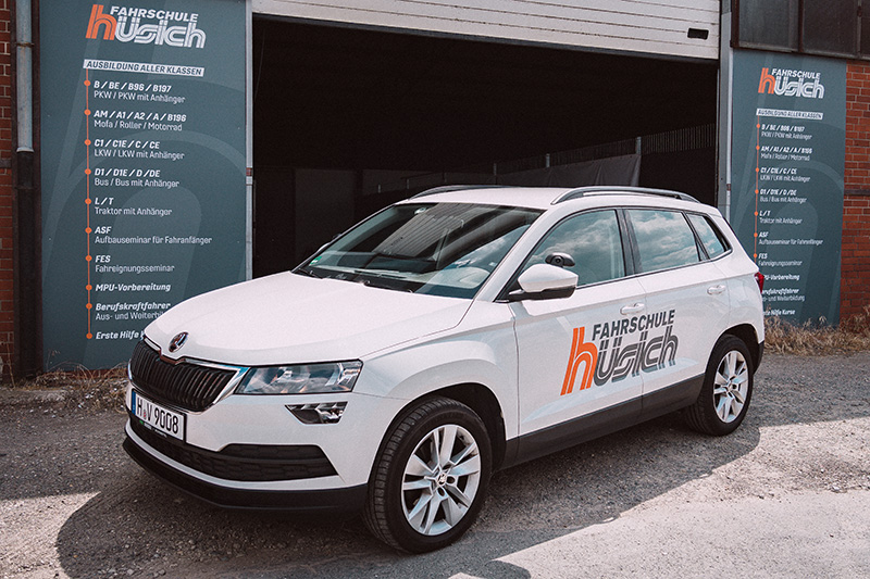 Ein weißer Skoda Karoq mit dem orangen Logo der Fahrschule Hüsich auf der Tür steht vor der Einfahrt des Gebäudes. Im Hintergrund sind zwei Schilder mit der Übersicht aller Führerscheinklassen zu sehen.