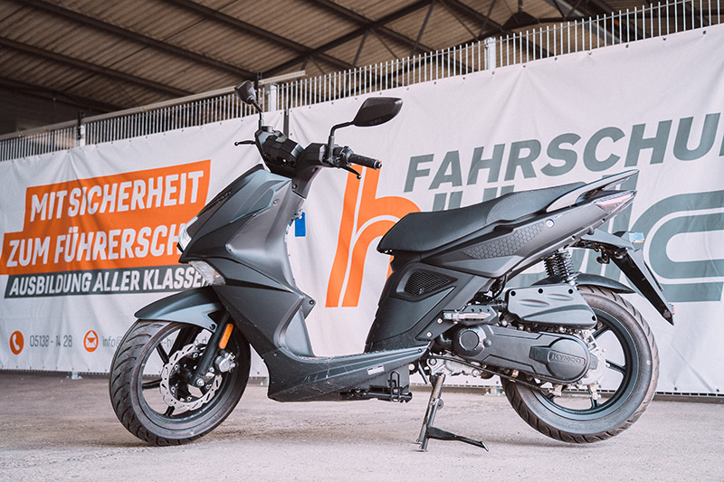 Ein matt-schwarzer Kymco Motorroller steht vor dem orange-weißen Werbebanner der Fahrschule Hüsich.