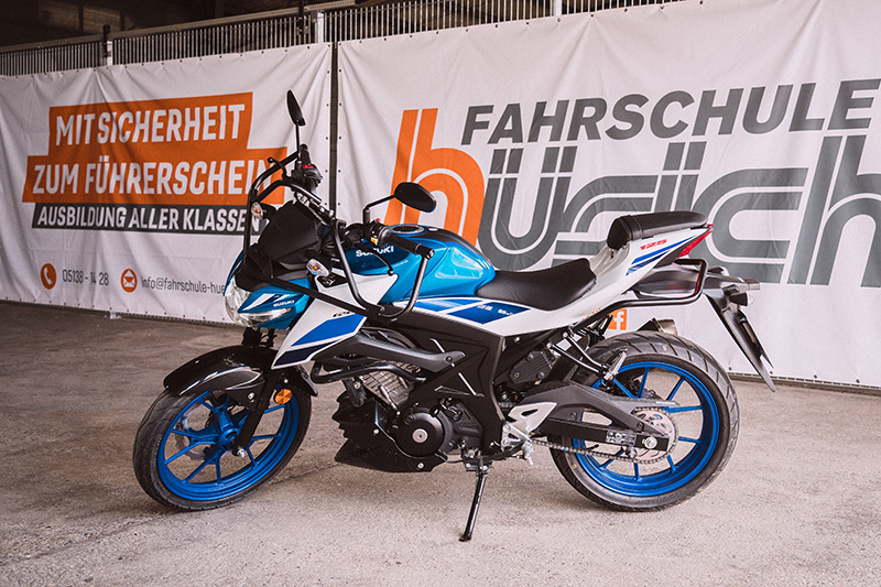 Ein blau-weißes Suzuki Motorrad steht vor einem Banner der Fahrschule Hüsich, auf dem 'Mit Sicherheit zum Führerschein' und 'Ausbildung aller Klassen' steht.