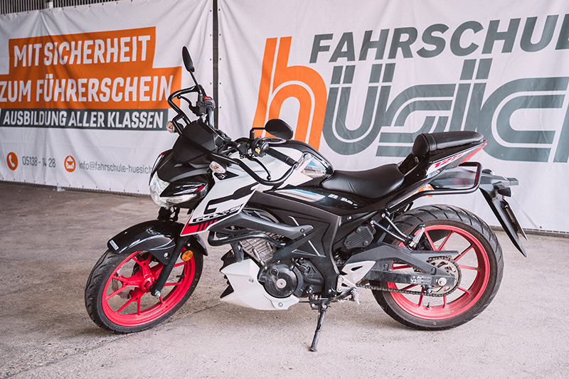 Ein schwarz-weißes Motorrad mit roten Felgen steht vor dem orange-weißen Banner der Fahrschule Hüsich, das 'Mit Sicherheit zum Führerschein' bewirbt.