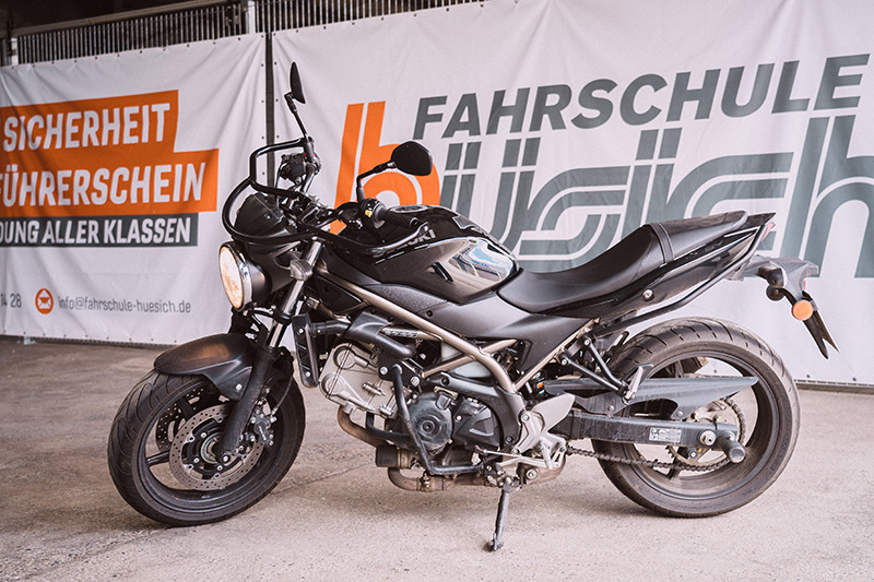 Ein schwarzes Motorrad steht vor einem orange-weißen Banner der Fahrschule Hüsich, das den Slogan 'Mit Sicherheit zum Führerschein' zeigt.