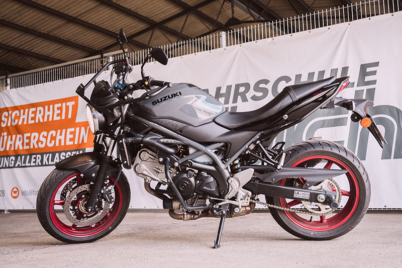 Ein mattgraues Suzuki Motorrad mit roten Felgen steht vor dem orange-weißen Banner der Fahrschule Hüsich.