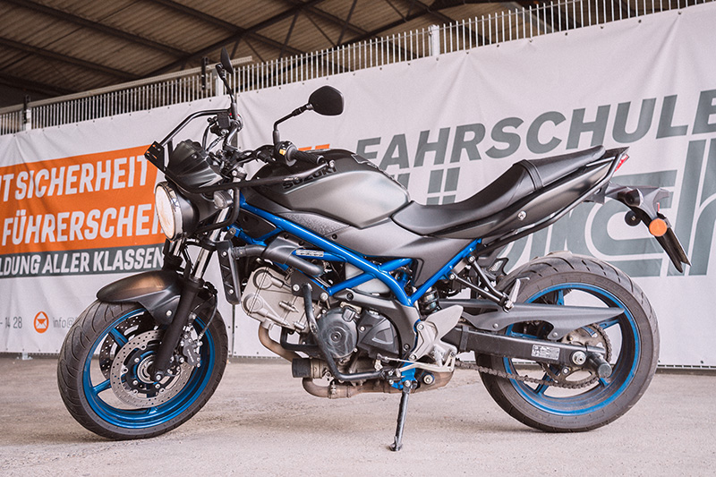 Ein mattgraues Suzuki Motorrad mit blauem Rahmen und blauen Felgen steht vor dem orange-weißen Banner der Fahrschule Hüsich.