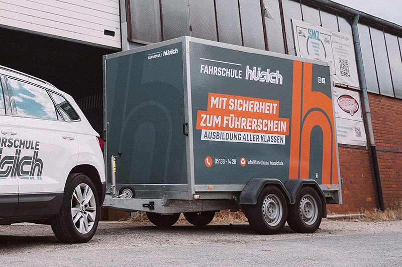 Nahaufnahme des dunkelgrauen Anhängers der Fahrschule Hüsich mit dem orangefarbenen Logo und dem Slogan 'Mit Sicherheit zum Führerschein' auf der Seite, angekoppelt an das weiße Fahrschulauto.
