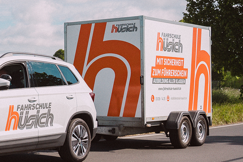 Heckansicht eines weißen Fahrschulautos (Skoda Karoq) der Fahrschule Hüsich, das einen Anhänger mit dem Logo und dem Slogan 'Mit Sicherheit zum Führerschein' auf der Straße zieht.