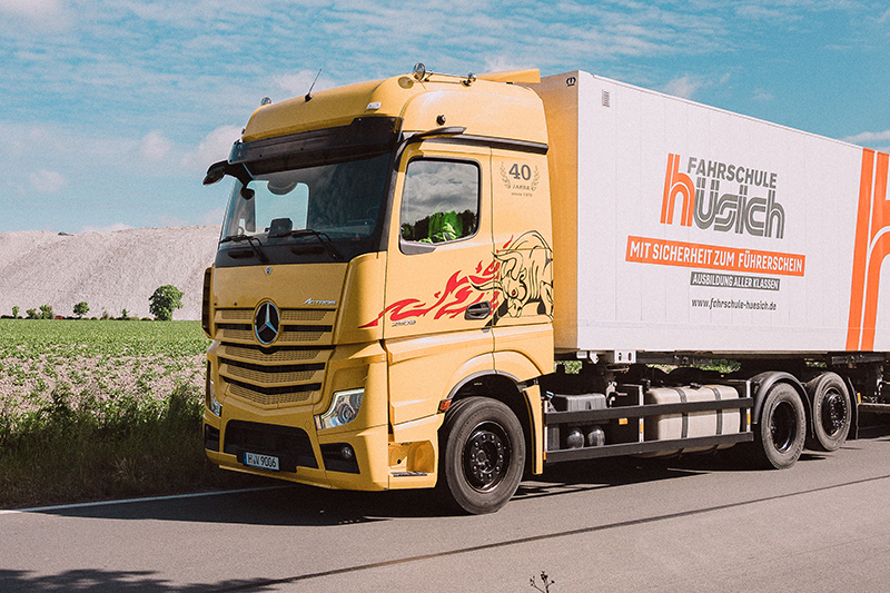 Nahaufnahme des gelben Mercedes-Benz Actros LKW der Fahrschule Hüsich mit Drachen-Branding auf der Fahrerkabine. Rechts ist der Anhänger mit dem Logo der Fahrschule zu sehen, im Hintergrund ein Salzberg.