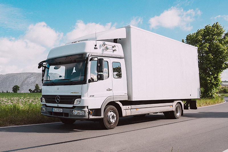 Ein weißer Mercedes-Benz Atego LKW mit Kofferaufbau fährt auf einer Straße. Im Hintergrund sind ein Feld, Bäume und ein großer weißer Salzberg sichtbar.