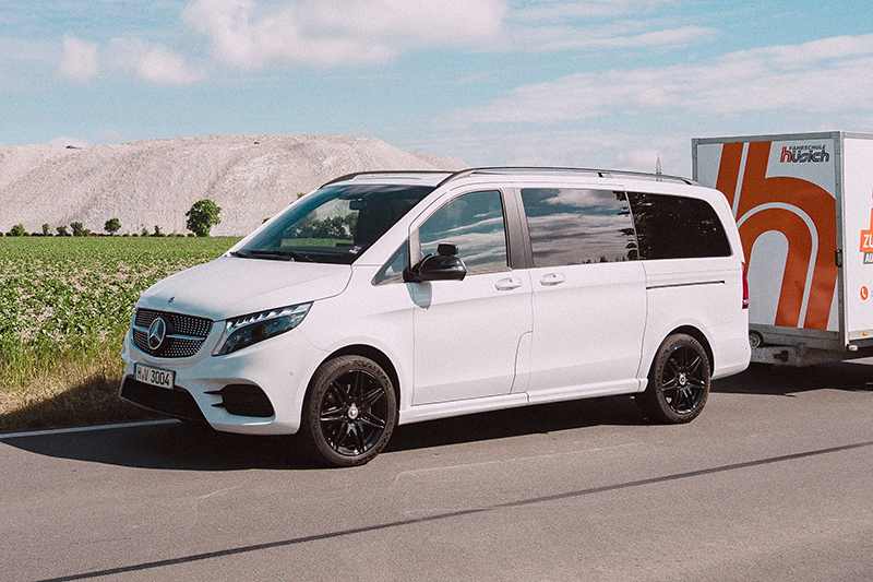Ein weißer Mercedes-Benz V-Klasse Van, gezogen von einem Anhänger mit dem Fahrschul Hüsich Logo, steht am Straßenrand. Im Hintergrund ist ein großer Salzberg sichtbar.