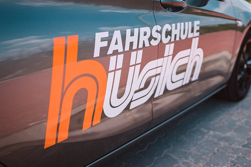 Nahaufnahme des großen Logos der Fahrschule Hüsich, mit dem orangen Buchstaben 'h' und dem weißen Schriftzug auf der Seitenverkleidung eines grauen Fahrschulautos.