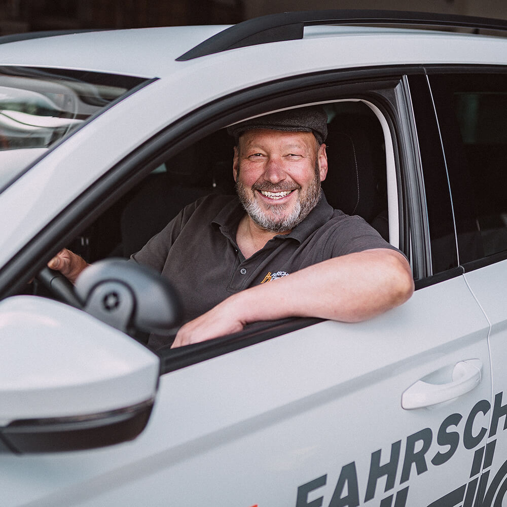 Ein lächelnder, bärtiger Mann mit Schiebermütze und einem Polohemd mit dem Logo der Fahrschule Hüsich sitzt am Steuer eines weißen Fahrschulautos.