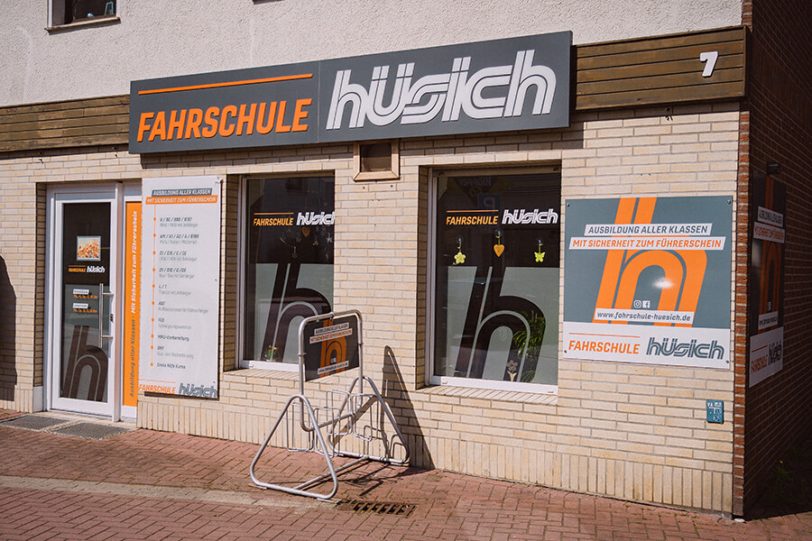 Außenansicht der Fahrschule Hüsich in einem hellen Backsteingebäude. Über der Eingangstür und den Fenstern befindet sich ein großes Schild mit dem Schriftzug „Fahrschule Hüsich“ in Orange und Weiß. Neben der Tür hängt eine Tafel mit den angebotenen Führerscheinklassen, vor dem Gebäude steht ein Fahrradständer.